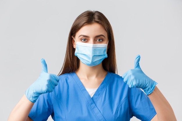 Covid-19, 바이러스, 건강, 의료 종사자 및 검역 개념을 예방합니다. 파란색 수술복, 의료 마스크, 장갑을 끼고 지지하는 여성 간호사나 의사의 클로즈업, 승인을 받은 엄지손가락
