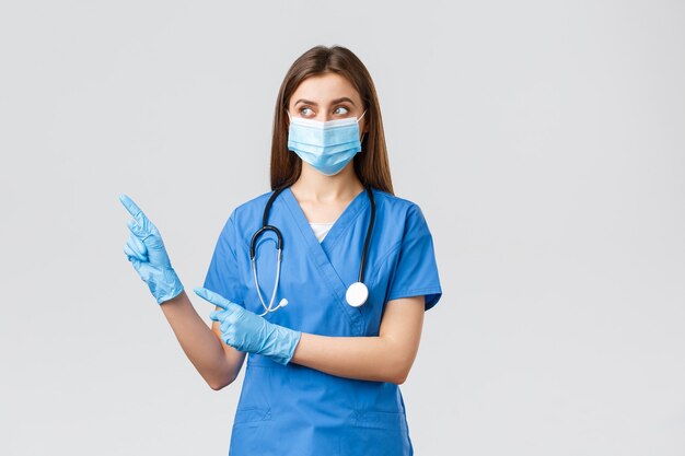 Covid-19, preventing virus, health, healthcare workers and quarantine concept. Doctor, female nurse in medical mask and blue scrubs, looking and pointing left at banner with coronavirus info