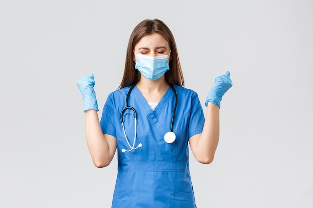 Covid-19, preventing virus, health, healthcare workers and quarantine concept. Encouraged happy female nurse or doctor in blue scrubs, celebrating, wear medical mask, chanting yes in success