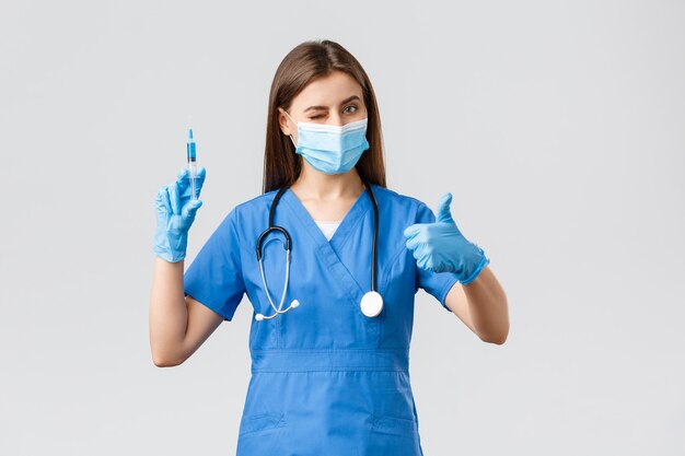 Covid-19, preventing virus, health, healthcare workers and quarantine concept. Friendly female nurse or doctor in blue scurbs, personal protective equipment, wink and assure need make shot of vaccine