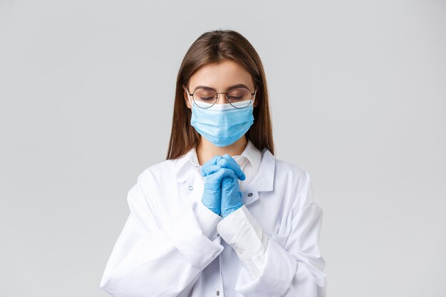 Covid-19, preventing virus, health, healthcare workers and quarantine concept. Hopeful doctor in scrubs and medical mask, personal protective equipment, praying with hands clasped together, hope