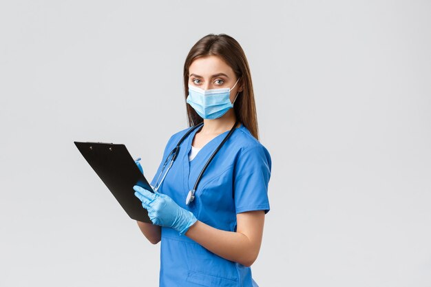 Covid-19, 바이러스, 건강, 의료 종사자 및 검역 개념을 예방합니다. 파란색 스크럽, 의료 마스크, 장갑을 끼고 클립보드를 사용하여 환자 정보를 기록하는 전문 여성 간호사 또는 의사