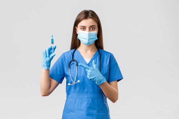 Covid-19, 바이러스, 건강, 의료 종사자 및 검역 개념을 예방합니다. 파란색 수술복을 입은 진지한 여성 간호사나 의사, 의료 마스크, 코로나바이러스 백신이 있는 주사기를 가리키는