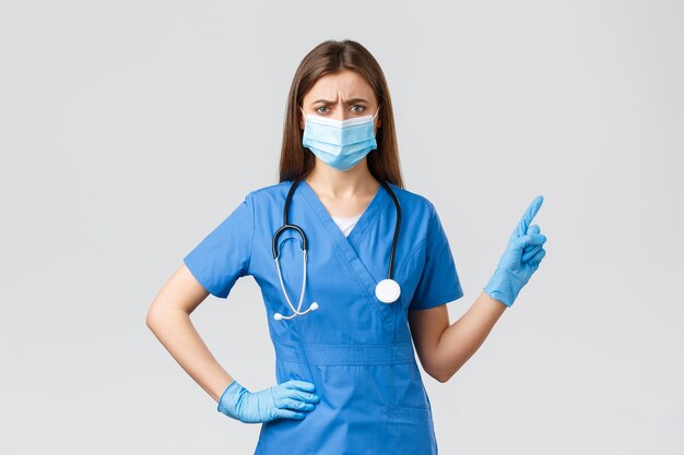 Covid-19, preventing virus, health, healthcare workers and quarantine concept. Skeptical or doubtful female nurse in blue scrubs and medical mask, gloves pointing right, frowning disbelief