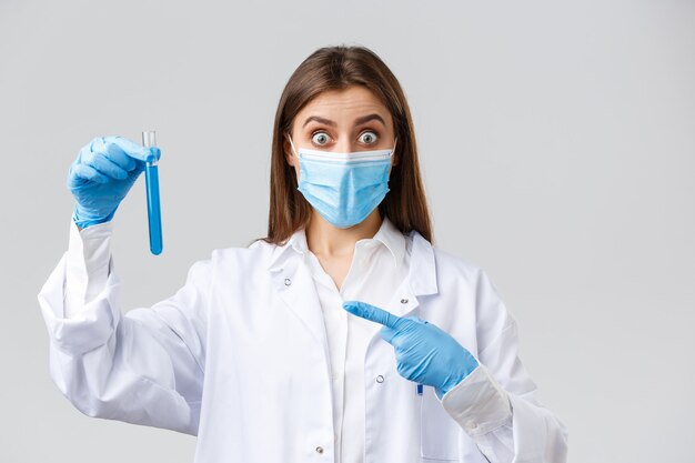 Covid-19, preventing virus, healthcare workers concept. Excited and surprised doctor in medical mask, gloves and scrubs pointing finger at test-tube coronavirus vaccine, impressive sample