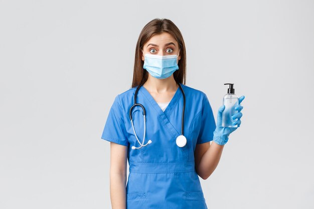 Covid-19, профилактика вируса, концепция медицинских работников. Удивленная медсестра или врач в синих скрабах, медицинской маске и перчатках объясняют важность дезинфицирующего средства для рук против коронавируса