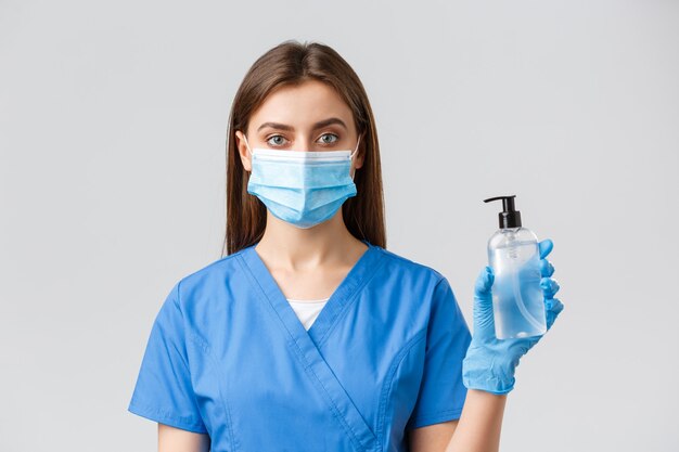 Covid-19, preventing virus, healthcare workers concept. Young pretty professional doctor, female nurse in medical mask and scrubs, showing hand sanitizer or soap to protect from coronavirus.