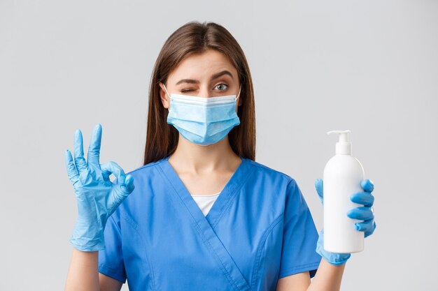 Covid-19, preventing virus, healthcare workers and quarantine concept. Confident cute female nurse or doctor in blue scrubs, medical mask and gloves, approve and recommend soap or hand sanitizer