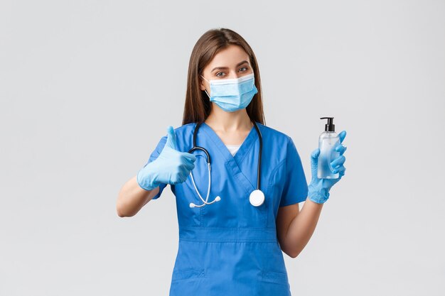 Covid-19, 바이러스, 의료 종사자 및 검역 개념을 예방합니다. 파란색 수술복을 입은 자신감 있는 여성 간호사 또는 의사, 의료 마스크 보호 장비, 엄지손가락으로 손 소독제 사용을 권장합니다