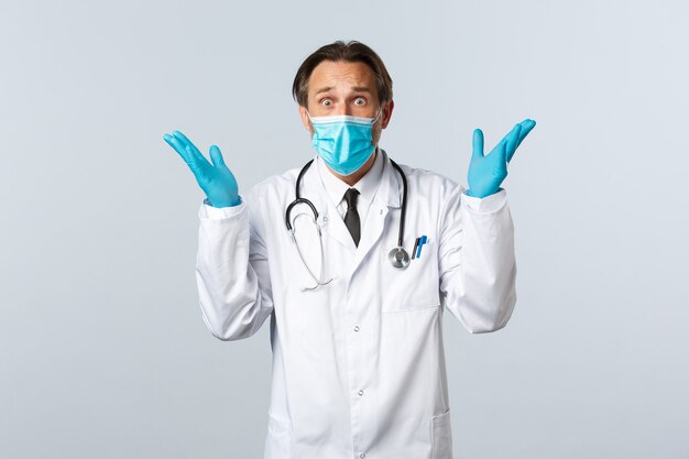 Covid-19, preventing virus, healthcare workers and vaccination concept. Alarmed worried doctor in medical mask and gloves, staring anxious with hands up distressed, panic over white background