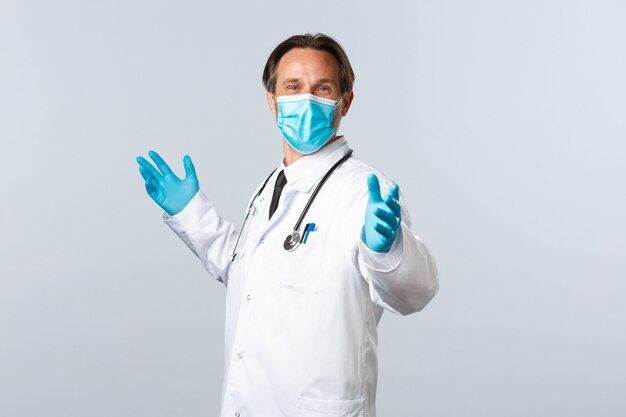 Covid-19, preventing virus, healthcare workers and vaccination concept. Cheerful glad doctor in medical mask and gloves, spread hands sideways relieved, celebrating good news, white background