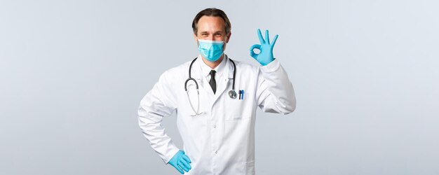 Covid-19, профилактика вируса, медицинские работники и концепция вакцинации. Врач клиники в медицинской маске и перчатках предоставляет услуги высочайшего качества, улыбаясь и показывая нормальный жест.