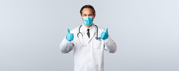 Covid-19, preventing virus, healthcare workers and vaccination concept. Confident handsome doctor in medical mask and gloves, show thumb-up, encourage or recommend clinic services