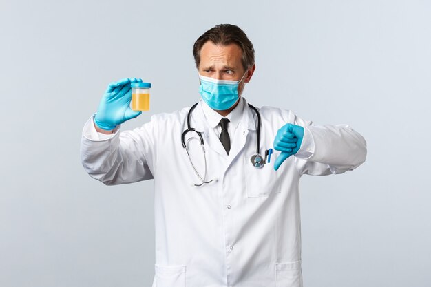 Covid-19, preventing virus, healthcare workers and vaccination concept. Disappointed doctor in medical mask and gloves show urine sample, thumbs-down have bad test result, white background