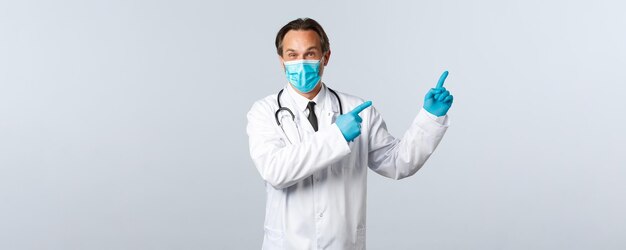Covid-19, профилактика вируса, медицинские работники и концепция вакцинации. Взволнованный и заинтригованный мужчина-врач в белом халате, медицинской маске и перчатках, укажите в правом верхнем углу, покажите рекламу клиники
