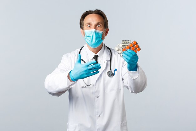 Covid-19, preventing virus, healthcare workers and vaccination concept. Excited and shocked doctor in medical mask and gloves react to new medication, showing pills, white background