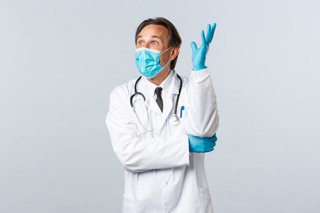 Covid-19, preventing virus, healthcare workers and vaccination concept. Excited upbeat male doctor in medical mask and gloves, raising hand up thoughtful, have idea, look amazed