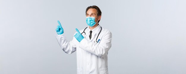 Covid-19, preventing virus, healthcare workers and vaccination concept. Middle-aged male doctor in white coat, medical mask and gloves looking upper left corner at pointing banner.