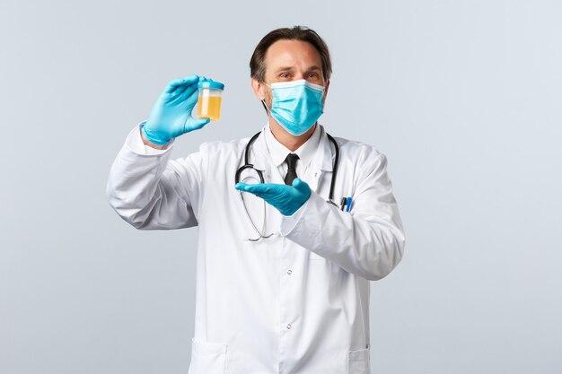 Covid-19, preventing virus, healthcare workers and vaccination concept. Pleased smiling doctor in medical mask and gloves show urine sample, satisfied with clean test result, white background