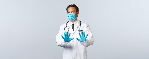 Covid-19, preventing virus, healthcare workers and vaccination concept. Reluctant or disgusted male doctor in medical mask and gloves, show refusal gesture, shaking hands in denial, white background