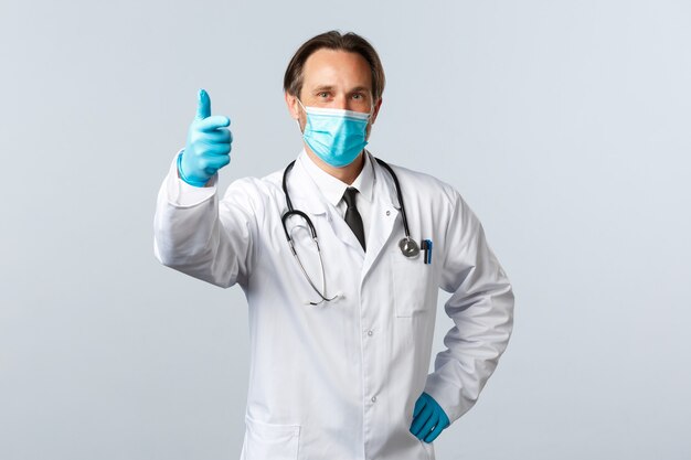 Covid-19, preventing virus, healthcare workers and vaccination concept. Satisfied doctor in medical mask and gloves, show thumb-up, well played gesture, pleased with advertisement promo