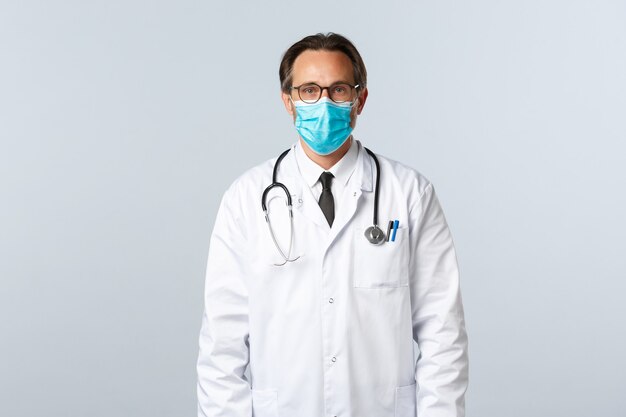 Covid-19, preventing virus, healthcare workers and vaccination concept. Serious confident male doctor in white coat and medical mask, wearing glasses, ready treat patient with influenza