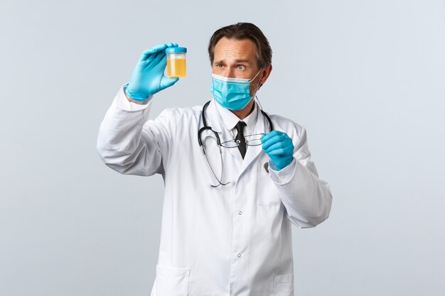 Covid-19, preventing virus, healthcare workers and vaccination concept. Serious-looking doctor in medical mask and gloves examine urine sample, look thoughtful and concentrated