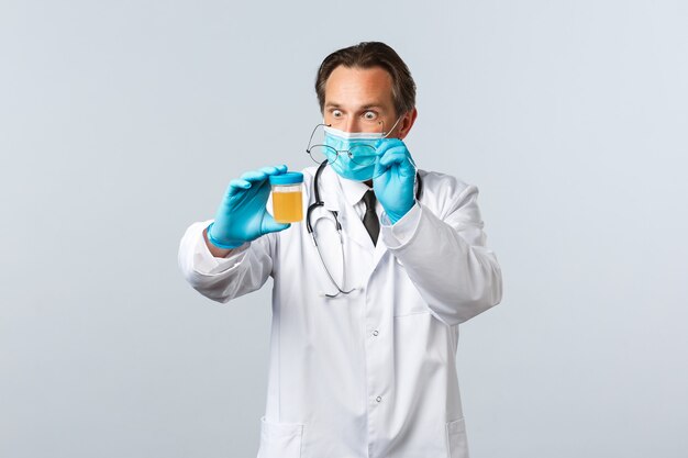 Covid-19, preventing virus, healthcare workers and vaccination concept. Shocked doctor in medical mask and gloves working in clinic lab, staring startled at urine sample