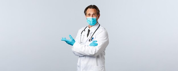 Covid-19, preventing virus, healthcare workers and vaccination concept. Skeptical confused doctor in medical mask and gloves pointing disappointed left, express frustration and disagreement.