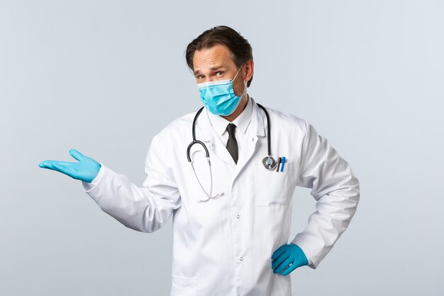 Covid-19, preventing virus, healthcare workers and vaccination concept. Skeptical male doctor in medical mask and gloves pointing left and staring camera displeased, give negative opinion