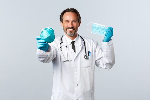 Covid-19, профилактика вируса, медицинские работники и концепция вакцинации. Улыбающийся харизматичный доктор в перчатках и белом халате рекомендует медицинский респиратор и маски, держится на социальной дистанции.