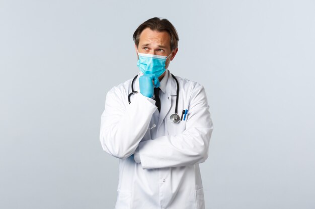 Free photo covid-19, preventing virus, healthcare workers and vaccination concept. thoughtful serious male doctor in medical mask and gloves thinking, making choice or decision, white background.