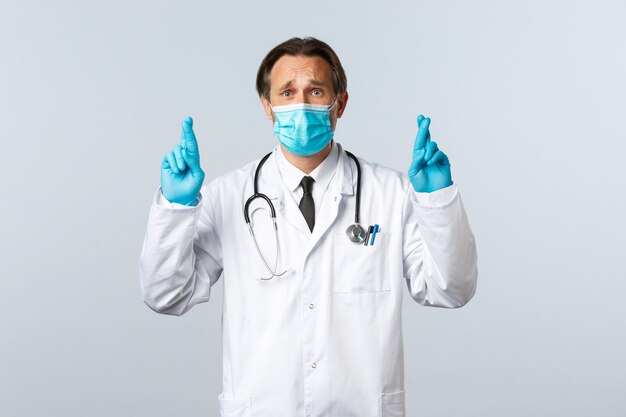 Covid-19, preventing virus, healthcare workers and vaccination concept. Troubled hopeful doctor in medical mask and gloves, cross fingers good luck, praying or making wish, white background