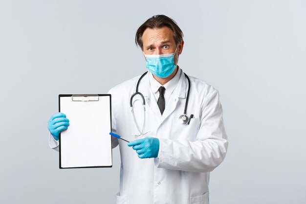 Covid-19, preventing virus, healthcare workers and vaccination concept. Worried and confused male doctor in medical mask and gloves, pointing at clipboard with information or diagnose