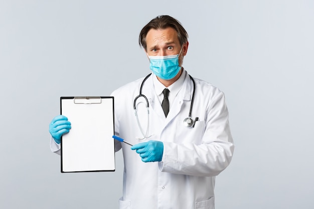 Covid-19, preventing virus, healthcare workers and vaccination concept. Worried and confused male doctor in medical mask and gloves, pointing at clipboard with information or diagnose