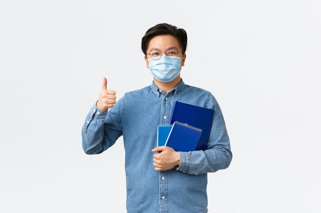 코비드-19, 바이러스 예방, 대학 개념에서의 사회적 거리두기. 쾌활한 아시아 남성 교사나 의료용 마스크를 쓴 교사는 공책과 학습 자료를 들고 엄지손가락을 치켜들고 흰색 배경을 보여줍니다.