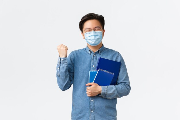 Covid-19, preventing virus, and social distancing at university concept. Rejoicing satisfied asian student passed exams, wearing medical mask, carry notebooks and fist pump, winning or triumphing.