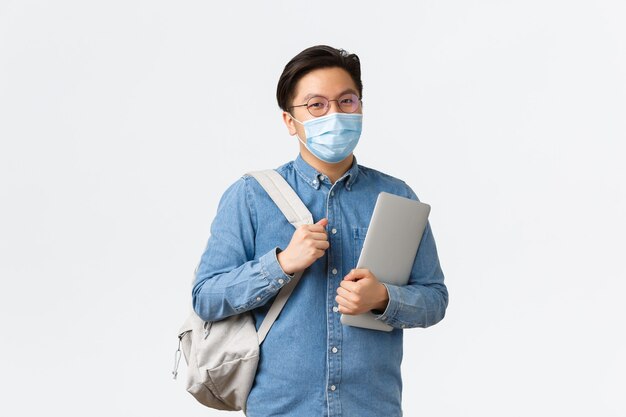 Covid-19, preventing virus, and social distancing at university concept. Smiling asian male student in medical mask going to lessons at college, holding laptop and backpack, standing white background.