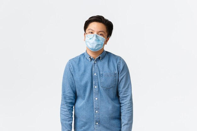 Covid-19, профилактика вируса и концепция социального дистанцирования на рабочем месте. Переутомленный и уставший азиатский служащий-мужчина выглядит неохотно, в медицинской маске, скучает, стоит на белом фоне