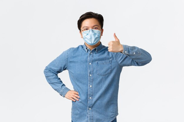 Covid-19, preventing virus, and social distancing at workplace concept. Supportive cheerful asian male entrepreneur in medical mask showing thumbs-up in approval, praise teamwork.