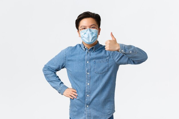 Covid-19、ウイルスの予防、職場での社会的距離の概念。承認で親指を立てて示す医療マスクの支持的な陽気なアジアの男性起業家は、チームワークを賞賛します。