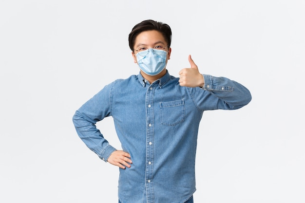 Covid-19, профилактика вируса и концепция социального дистанцирования на рабочем месте. Поддерживающий жизнерадостный азиатский предприниматель мужского пола в медицинской маске показывает большие пальцы в знак одобрения, хвалит командную работу.