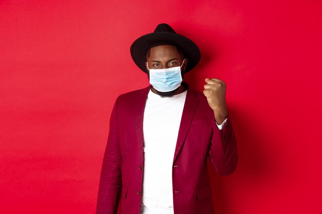 코비드-19, 검역 및 휴일 개념. 주먹을 꽉 쥐고 승리를 기뻐하고, 목표를 달성하고, 코로나바이러스의 의료용 마스크를 쓰고 있는 쾌활한 아프리카계 미국인 남자