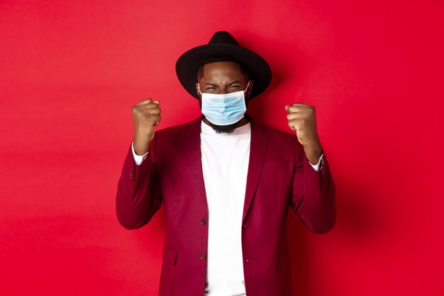 코비드-19, 검역 및 휴일 개념. 주먹을 꽉 쥐고 승리를 기뻐하고, 목표를 달성하고, 코로나바이러스의 의료용 마스크를 쓰고 있는 쾌활한 아프리카계 미국인 남자