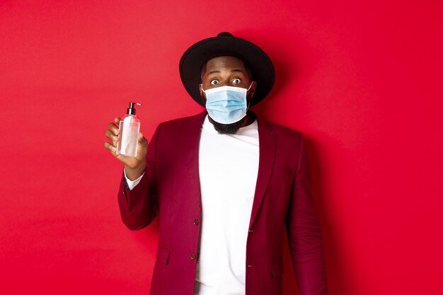 Covid-19, quarantine and holidays concept. Young african american man in medical mask and stylish outfit showing hand sanitizer, recommending to use antiseptic