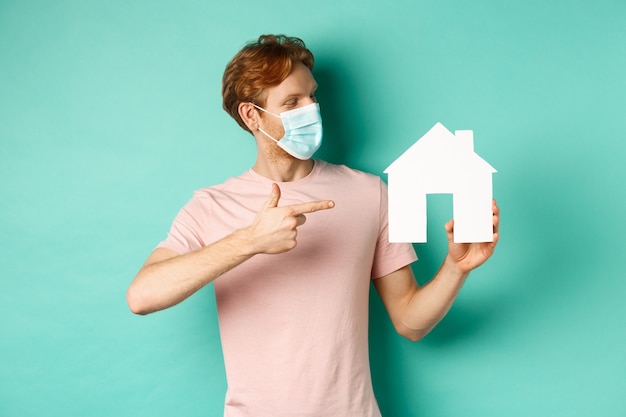 Covid-19 and real estate concept. Redhead guy in medical mask pointing and looking at paper house cutout, standing over turquoise background.