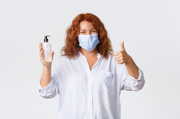 Covid-19 social distancing, coronavirus preventing measures and people concept. Smiling cute middle-aged redhead lady recommend hand sanitizer, showing thumbs-up and wearing medical mask.