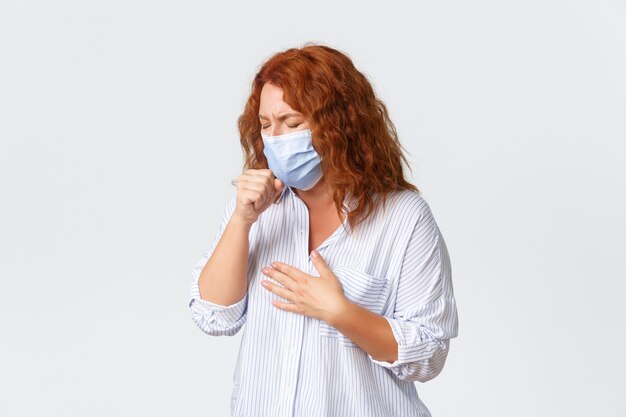 Covid-19 social distancing, coronavirus self-quarantine and people concept. Sick middle-aged redhead woman coughing, wearing medical mask, having sour throat, disease symptoms, caught influenza.