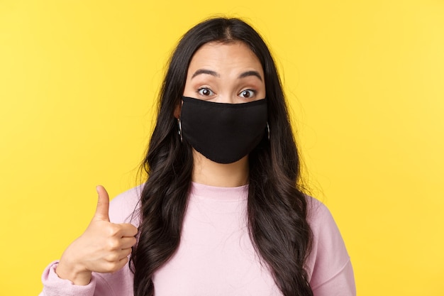Covid-19, social-distancing lifestyle, prevent virus spread concept. Close-up of enthusiastic asian girl in face mask, look surprised and amused, showing thumbs-up in approval, yellow background