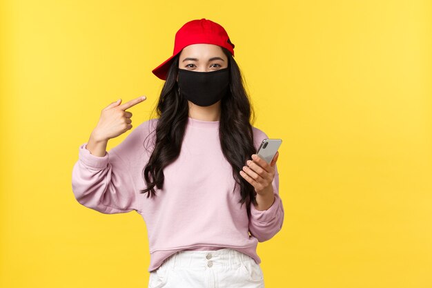 Covid-19, social-distancing lifestyle, prevent virus spread concept. Smiling cute asian girl pointing at face mask, asking protect health during coronavirus, holding mobile phone, yellow background